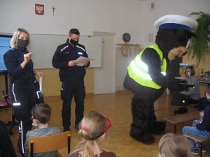 Maskotka podkarpackiej Policji - miś POPO podczas spotkania z dziećmi. 
Osoba ubrana w kostium misia w stroju policjanta wręcza dzieciom opaski odblaskowe. Na drugim planie umundurowani policjantka i policjant.