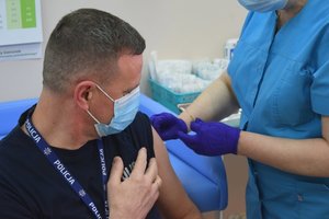 Mł.insp. Stanisław Sekuła Zastępca Komendanta Wojewódzkiego Policji w Rzeszowie podczas szczepienia. Zdjęcie wykonane w gabinecie zabiegowym szpitala MSWiA w Rzeszowie. Komendant ubrany w granatowy t-shirt z białymi napisami POLICJA, na szyi granatową smycz z napisami POLICJA, na twarzy założona maseczka. Po prawej pielęgniarka ubrana w niebieski fartuch.