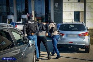w tle budynek prokuratury, na placu dwa samochody osobowe pomiędzy nimi dwóch mężczyzn w kamizelce z napisem policja prowadzi człowieka