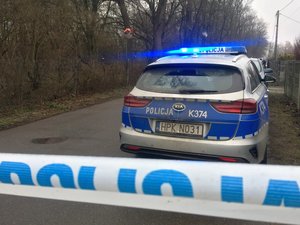 Na pierwszym planie biała taśma z niebieskim napisem POLICJA, za taśmą oznakowany radiowóz policyjny z włączonymi sygnałami świetlnymi stojący po prawej stronie ulicy, przy ogrodzeniu posesji.