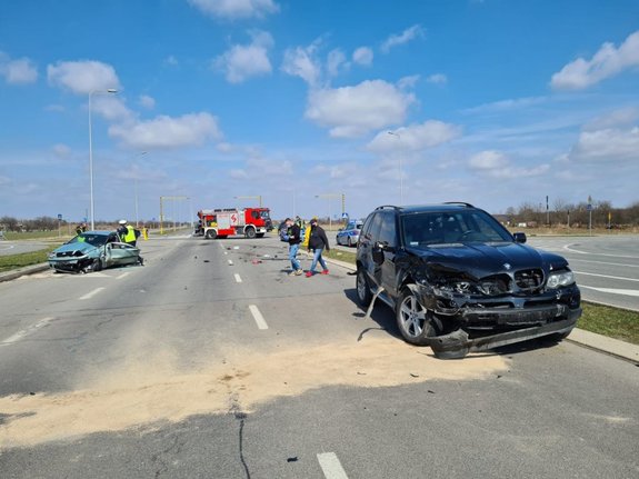 Zdjęcie kolorowe przedstawia odcinek obwodnicy przemyskiej. Droga trzy pasmowa z lewej strony zdjęcia widoczny samochód osobowy marki BMW po wypadku (uszkodzony) koloru granatowego tuz za samochodem widoczni dwaj mężczyźni ubrani w spodnie jeans oraz ciemne bluzy. W oddali na środku fotografii widoczny jest pojazd Straży pożarnej. Po stronie lewej  stoi uszkodzony pojazd marki Fiat Punto koloru jasno niebieskiego przy którym stoi 2-ch policjantów ruchu drogowego ubranych w mundury granatowe oraz kamizelki odblaskowe.