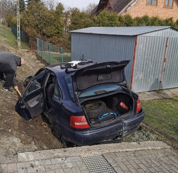 Zdjęcie pora dzienna przedstawia pojazd m-ki tył samochodu audi granatowe który jest  w rowie. Pojazd po kolizji. Z prawej strony pojazdy widoczny jest garaż blaszany  oraz za drzewani wierzchołek domu piętrowego zbudowanego  z cegły. Z lewej strony pojazdu widoczny mężczyzna  , ubrany w szara bluzę z kapturem o niebieskim kolorze oraz spodnie ciemne. Na nogach ma obuwie sportowe w kolorze niebieskim z białą podeszwa oraz na zewnętrznej stronie buta widnieje logo firmowe tz „łyżwa” w kolorze białym Mężczyzna trzyma w ręce łopatę i próbuje odkopać koło z samochodu