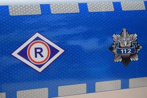 Na zdjęciu widoczny fragment drzwi policyjnego radiowozu. Na niebieskim tle po lewej symbol pionu ruchu drogowego, po prawej logo policji