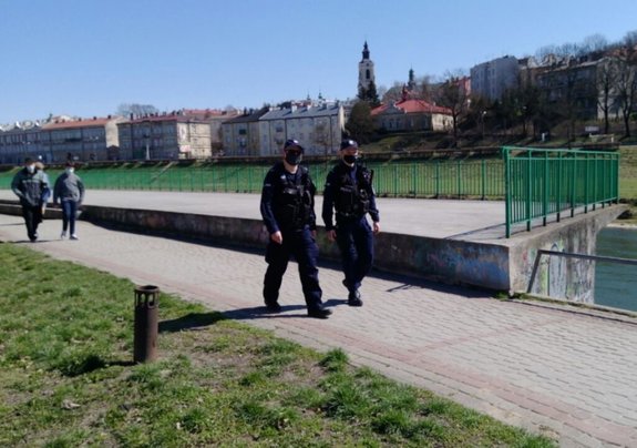 1.	Zdjęcie kolorowe przedstawia fragment deptana przy rzece San w Przemyślu. Na pierwszym planie fotografii widoczni dwaj policjanci, ubrani w granatowe mundury oraz kamizelki taktyczne. Policjanci w patrolu pieszym. Za policjantami widoczne sa trzy osoby spacerowicze . W tle fotografii widoczne sa stare kamienice Przemyśla.