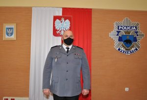 2.	Zdjęcie kolorowe przedstawia aulę Komendy Miejskiej Policji w Przemyślu. Na zdjęciu widoczny jest Komendant Miejskiej Policji w Przemyślu kom. Janusz Kiszka . Ubrany w strój galowy czarne spodnie, czarne buty, oraz popielata gabardyne. Za komendantem widoczna ściana na której jest zawieszona flaga Polski w kolorze biało czerwonej na której znajduje się na środku godło Polski (Orzeł) z prawej strony  flagi umieszczone jest logo Policji a z lewej strony zawieszony jest herb Miasta Przemyśla