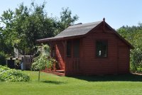 Zdjęcie kolorowe przedstawia mały domek letniskowy w kolorze brązowym, który stoi na działce ogródków działkowych. Za domem widoczne są drzewa. Na przednim planie widoczny trawnik