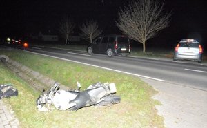 Zdjęcie przedstawia drogę, miejsce wypadku drogowego. Po lewej stronie na poboczu trawiastym znajduje się uszkodzony motocykl. Po prawej stronie jezdni widać radiowóz oraz samochód biorący udział w zdarzeniu.