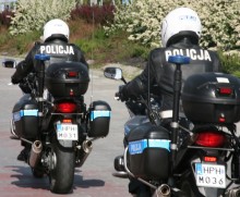 Zdjęcie kolorowe przedstawia drogę po której poruszają się dwa motory policyjne jadą jeden za drugim. Na motocyklach widoczni dwaj policjanci ubrani w mundury przeznaczone do jazdy na motocyklu. Na głowach maja białe kaski a na skórzanych kurtkach koloru czarnego białe napisy „POLICJA”