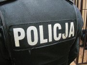 Zdjęcie kolorowe przedstawia plecy policjanta stojącego tyłem, ubranego w kamizelkę taktyczna koloru czarnego na środku której jest napis „POLICJA” w białym kolorze