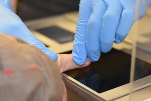 na zdjęciu dłonie w niebieskich rękawiczkach przyciskają palec do czarnej podkładki z tuszem