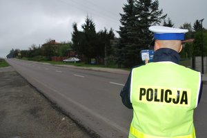 Zdjęcie kolorowe przedstawia policjanta wydziału ruchu drogowego który stoi tyłem w reku trzyma urządzenie do mierzenia prędkości skierowane na pojazdy. Policjant ubrany jest w granatowy mundur na którym nałożoną ma kamizelkę odblaskową koloru żółtego z napisem „POLICJA” w kolorze czarnym. W tle zdjęcia widoczna ulica jednopasmowa.