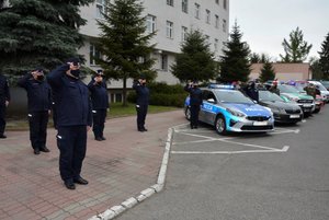 3. Zdjęcie kolorowe przedstawia plac Komendy Miejskiej Policji w Przemyślu . W tle widoczna jest część budynku komendy, przed budynkiem widoczne są zaparkowane pojazdy służb mundurowych oznakowane : policja, straż więzienna,straż graniczna , straż pożarna. Z lewej strony widoczni są policjanci ubrani w granatowe mundury  którzy trzymają, prawą rękę przy czole, oddają cześć poległemu Policjantowi . Przed radiowozami widoczni f-sze służb którzy także  trzymają prawą rękę przy czole, oddają cześć poległemu Policjantowi