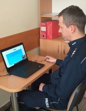 Zdjęcie przedstawia umundurowanego policjanta siedzącego przed komputerem i prowadzącego zdalne szkolenie dla dyrektorów szkół i nauczycieli pt. „Hejt i inne zagrożenia” z wykorzystaniem prezentacji multimedialnej.