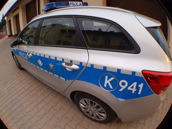 Oznakowany radiowóz policyjny koloru srebrnego z niebieskim pasem na boku. Kadr zniekształcony przez wykonanie zdjęcie obiektywem szerokokątnym.