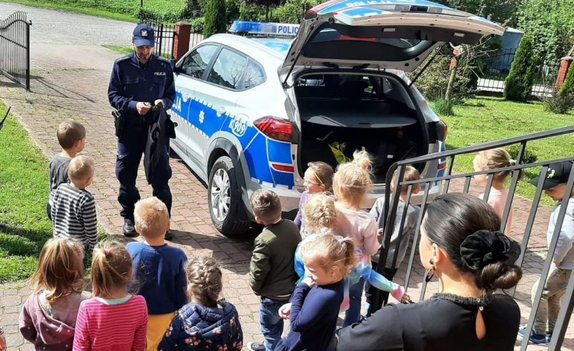 Plac przy przedszkolu. Na zdjęciu umundurowany policjant stoi przy radiowozie i pokazuje dzieciom wyposażenie do służby.