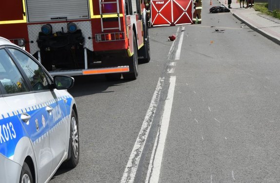 Miejsce zdarzenia. Po lewej stronie radiowóz, za nim wóz strażacki i parawan ochronny z napisem straż. Po prawej stronie na jezdni znajduje się  motorower.