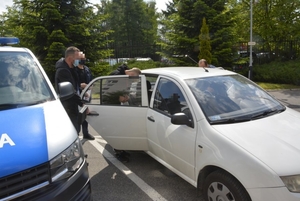 4.	Zdjęcie kolorowe przedstawia parking KMP w Przemyślu na zdjęciu z prawej strony widoczny jest pojazd nieoznakowany policyjny koloru białego do którego przez tylne drzwi od strony pasażera wprowadzony jest zatrzymany mężczyzna a obok niego stoi dwóch nieumundurowanych policjantów. Po lewej stronie zdjęcia częściowo widoczny jest pojazd policyjny oznakowany. W oddali widoczny jest parking samochodowy