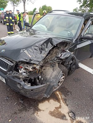 Zdjęcie kolorowe przedstawia miejsce zdarzenia drogowego. Na pierwszym planie widoczne uszkodzone auto marki BMW koloru ciemnego na drugim planie zdjęcia z lewej strony widoczni sa strażacy, policjant ruchu drogowego oraz trzy osoby cywilne.