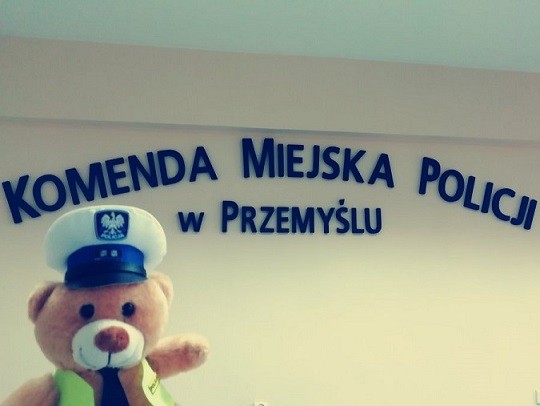 Zdjecie kolorowe przedstawia misia policyjnego a w tle widoczny napis &quot;KOMENDA MIEJSKA POLICJI W PRZEMYŚLA &quot; W NIEBIESKIM KOLORZE