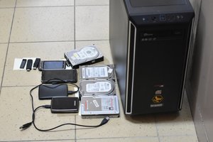 Na podłodze, patrząc od prawej strony czarna obudowa jednostki centralnej komputera, w środkowej części leżą cztery dyski zewnętrzne, obok nich, po lewej stronie cztery telefony komórkowe