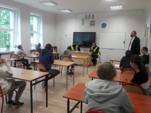 w sali lekcyjnej siedzi kilkunastu uczniów podczas testu wiedzy z przepisów ruchu drogowego, w komisji, za biurkiem siedzi 3 umundurowanych policjantów