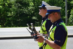 Dwaj policjanci ruchu drogowego, umundurowani i w odblaskowych kamizelkach, trzymają w rękach panel sterowania dronem.