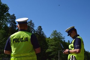Dwaj policjanci ruchu drogowego, umundurowani i w odblaskowych kamizelkach, trzymają w rękach panel sterowania dronem. W górze widoczny dron.