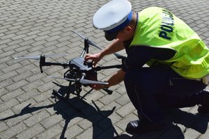 Policjant ruchu drogowego w odblaskowej kamizelce podłącza akumulator do drona stojącego na chodniku.