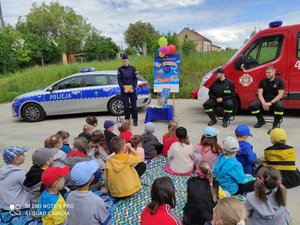 na zdjęciu przedszkolaki wysłuchują prelekcji policjantki oraz strażaków. w tle policyjny radiowóz oraz wóz strażacki