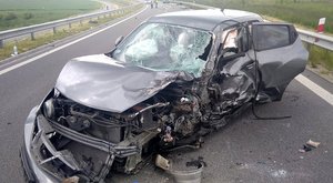 Rozbity samochód nissan stoi na środku drogi. Ma zniszczony pas przedni, wyrwany zderzak, lewy reflektor i lewy przedni błotnik, rozbitą szybę czołową i zniszczone drzwi kierowcy.