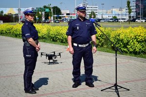 Policjanci ruchu drogowego podczas briefingu prasowego na parkingu przy Parku Papieskim, stoją przed mikrofonem. Po lewej policjantka, po prawej policjant.