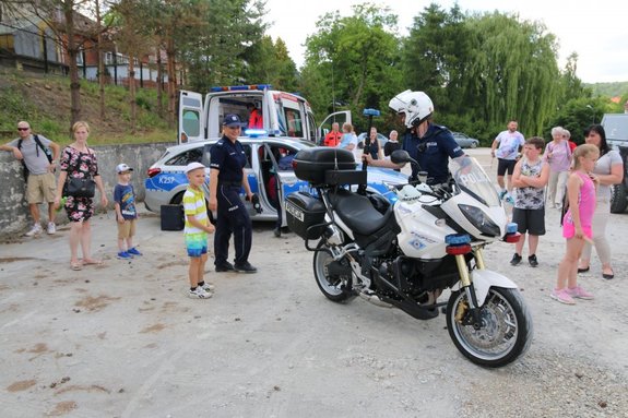 Zdjęcie kolorowe przedstawia piknik rodzinny w Przemyślu na Osiedlu Kazanów. 
W tle fotografii widoczny jest policyjny radiowóz oznakowany oraz pojazd pogotowia ratunkowego . Wokoło pojazdów widoczni są uczestnicy pikniku oraz umundurowana policjantka. Na przednim planie widoczny jest policjant ruchu drogowego który prowadzi motocykl policyjny.