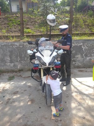 . Zdjęcie kolorowe przedstawia piknik rodzinny w Przemyślu na Osiedlu Kazanów. 
Na fotografii widoczny jest motocykl policyjny na którym siedzi mały chłopiec uczestnik pikniku  a obok niego stoi policjant ruchu drogowego. Na przednim planie zdjęcia widoczny jest drugi chłopiec który rękoma trzyma przednie koło motoru. Chłopiec odwrócony jest tyłem do zdjęcia .