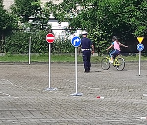 Uczestnik egzaminu na kartę rowerową jadący po wyznaczonym torze, na którym znajdują się różne znaki drogowe. Za nim oceniający go policjant.