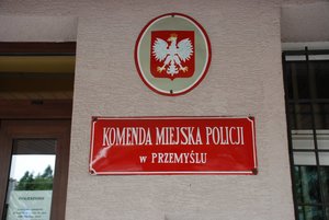 zdjęcie przedstawia front budynku Komendy Miejskiej Policji w Przemyślu. Na środku fotografii widoczna ściana na której są zawieszone dwie tabliczki, górna tablica przedstawia godło Polski ( orła w koronie), pod godłem umieszczona tabliczka w czerwonym kolorze z białymi napisami „KOMENDA MIEJSKA POLICJI w PRZEMYŚLU”