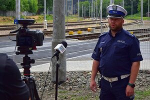 Policjant ruchu drogowego stoi przed mikrofonem. W tle przejazxd kolejowy.