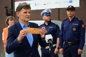 Uczestnicy briefingu na przejeździe kolejowym. Mężczyzna w garniturze na pierwszym planie pokazuje żółtą naklejkę. W tle zaproszeni goście, m.in. policjant.