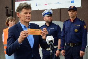 Uczestnicy briefingu na przejeździe kolejowym. Mężczyzna w garniturze na pierwszym planie pokazuje żółtą naklejkę. W tle zaproszeni goście, m.in. policjant.