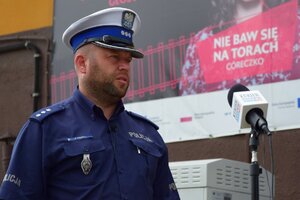 Policjant ruchu drogowego stoi przed mikrofonem. W tle napis "Nie baw się na torach córeczko".