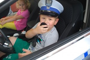 Chłopiec w policyjnej czapce policjanta ruchu drogowego, siedzący w radiowozie. W ręku trzyma manetkę radiostacji,