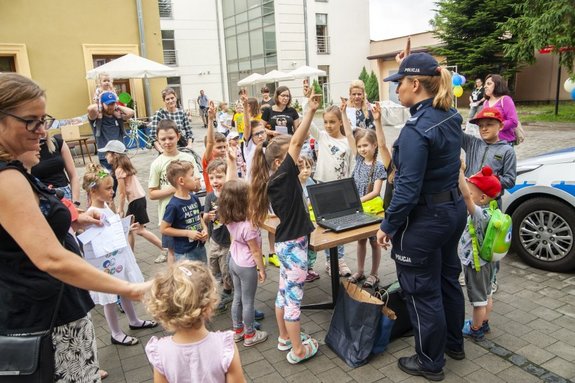 Zdjęcie kolorowe przedstawia plac parkingowy Biblioteki Publicznej w Przemyślu, miejsce spotkania dzieci z policjantem. Na zdjęciu jest widoczna policjantka przed która stoi gromadka dzieci biorąca udział w zajęciach na temat bezpiecznych wakacji . w tle widać budynek biblioteki publicznej