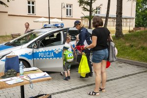 Zdjęcie kolorowe przedstawia plac parkingowy Biblioteki Publicznej w Przemyślu, miejsce spotkania dzieci z policjantem. Po prawej stroi zdjęcia widoczna jest policjantka która stoi przed stolikiem a w ręku trzyma kamizelkę odblaskową do niej podchodzi mała dziewczynka ubrana w jasną sukienkę. Po lewej stronie zdjęcia widoczni są ludzie którzy stoją na placu. W tle widoczny jest budynek biblioteki publicznej w Przemyślu.