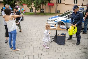 Zdjęie kolorowe przedstawia plac parkingowy Biblioteki Publicznej w Przemyślu, miejsce spotkania dzieci z policjantem. Na pierwszym planie fotografii widoczny jest radiowóz oznakowany policyjny przy którym stoi policjantka wraz z trójka dzieci. Na przednim planie widoczny jest stolik na którym znajdują się przedmioty , tarcza do zatrzymywania pojazdów torba koloru granatowego  oraz kartki do kolorowania, obok stoi kobieta ubrana w krótkie spodenki koloru niebieskiego oraz czarna koszulkę. Kobieta obrócona jest tyłem.