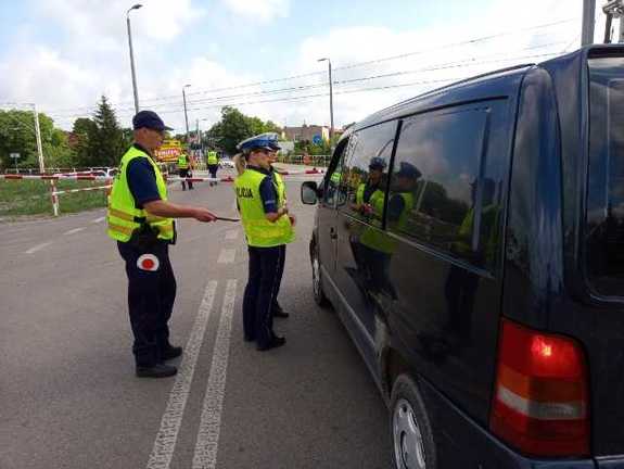 Na zdjęciu znajduje się pojazd osobowy przed przejazdem kolejowym z opuszczonymi rogatkami i dwoje umundurowanych policjantów w porze dziennej