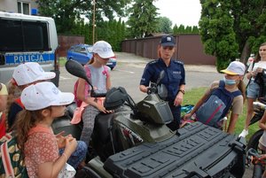 Zdjęcie kolorowe przedstawia przedstawia parking samochodowy Komendy Miejskiej Policji w Przemyślu. Po prawej stronie zdjęcia widoczna jest policjantka ubrana w granatowy mundur  a obok niej qład policyjny .Wokoło pojazdu oraz policjantki stoją dzieci które ogładają sprzęt policyjny. W oddali po lewej stronie  widać częściowo samochód oznakowany policyjny m-ki DUKATO. Za policjantką w tle widoczne są garaże