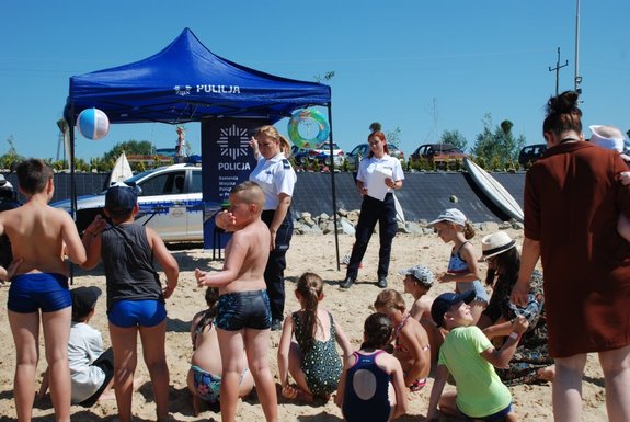 . Zdjęcie kolorowe przedstawia plażę w Ostrowie . Zdjęcie przedstawia grupę dzieci biorących udział w spotkaniu z policjantka  ubrana w granatowe spodnie oraz biała koszulkę która stoi przed namiotem policyjnym w kolorze . Policjantka jedna ręką wskazuje na namiot (ręka jest unieniona ku górze). Za namiotem widoczny jest radiowóz m-kia po lewej stronie zdjęcia w prawym rogu stoi druga policjantka ubrana w granatowe spodnie oraz biała koszulkę .