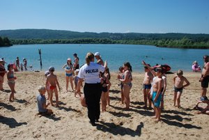 . Zdjęcie kolorowe przedstawia plażę w Ostrowie z widokiem na zalew wodny. W tle widoczny zalew a na piasku przed wodą widoczna jest grupa dzieci która stoi twarzami do policjantki która przekazuje dzieciom wiadomości na temat bezpieczeństwa nad wodą. Policjantka ubrana jest w granatowe spodnie oraz biała koszulkę z niebieskim napisem POLICJA na środku.