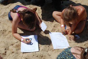 Na zdjęciu widoczne sa dwie dziewczynki które na piasku wypełniają test wiedzy o bezpieczeństwie nad wodą .