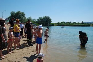 Zdjęcie przedstawia plażę w miejscowości Ostrów k. Przemyśla nad brzegiem której stoi grupa osób oglądająca wejście do wody nurka SPS –Przemyśl. Nurek jest na linie i tyłem wchodzi do wody w celach pokazowych.