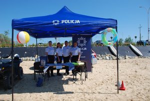 1.	Zdjęcie kolorowe przedstawia plażę w Ostrowie. Na zdjęciu widoczni są 4 policjanci (3 policjantki oraz 1 policjant) umundurowani w granatowe spodnie oraz białe koszulki z napisem Policja z lewej strony. Policjanci stoją pod namiotem przed nimi jest stoisko z różnymi ulotkami , słodyczami i nagrodami na konkurs.